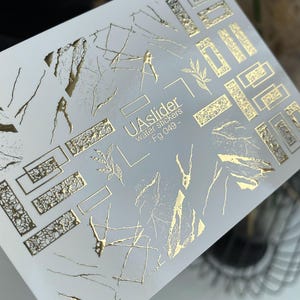 May include: A sheet of water transfer nail art stickers. The stickers feature gold geometric shapes, leaf designs, and abstract lines on a white background. The text "UAslider water stickers - Fg 049 -" is printed on the sheet.