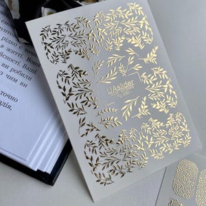 Może przedstawiać: Arkusz naklejek do zdobienia paznokci ze złotymi wzorami liści. Naklejki zawierają różne wzory liści i gałęzi, z nadrukowanym tekstem "UAslider water stickers Fg 030". Naklejki są na białym tle.