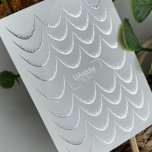 Pode incluir: Uma folha de autocolantes de água para nail art com um padrão de ondas prateadas repetidas. O design tem um efeito texturizado e brilhante. O texto "UAslider Water stickers - Fs 281" está impresso.