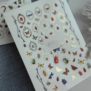 Może przedstawiać: Arkusz naklejek do zdobienia paznokci z transferem wodnym przedstawiających różne owady, w tym motyle, chrząszcze i ważki. Naklejki mają złote i kolorowe akcenty, a niektóre owady są oprawione w ozdobne wzory. Widoczny jest tekst "UAslider".