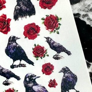 May include: Sheet of stickers featuring illustrations of black crows and red roses. The crows are depicted in various poses, some perched and others in flight. The roses have a glittery effect, adding a touch of sparkle.
