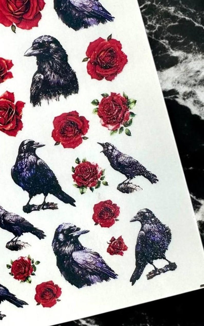 May include: Sheet of stickers featuring illustrations of black crows and red roses. The crows are depicted in various poses, some perched and others in flight. The roses have a glittery effect, adding a touch of sparkle.