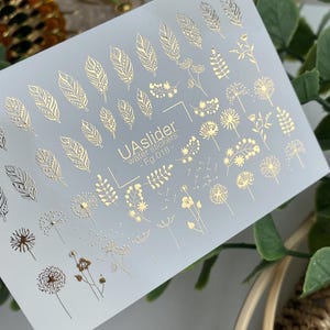 May include: A sheet of water transfer nail art stickers featuring gold foil designs of feathers, dandelions, and other floral elements. The text "UAslider water stickers - Fg 018 -" is printed on the sheet.