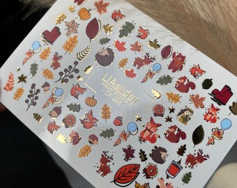 Puede incluir: Una lámina de pegatinas de transferencia de agua para uñas con diseños otoñales. Las pegatinas incluyen hojas, calabazas, zorros, erizos y bellotas en tonos naranja, marrón y dorado. El texto "UA slider water stickers - CF 288" está impreso.