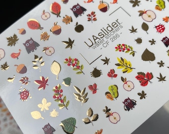 Puede incluir: Hoja de pegatinas de transferencia de agua para uñas con diseños de temática otoñal. Las pegatinas presentan hojas, manzanas, bellotas, búhos y mariquitas doradas, marrones, rojas y verdes. El texto "UAslider water stickers - CF 286" está impreso.