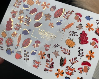 Puede incluir: Una lámina de pegatinas para uñas con transferencia de agua con temática otoñal. Las pegatinas presentan ilustraciones coloridas de hojas, calabazas, setas, zorros y otros diseños relacionados con el otoño. El texto "UA slider water stickers - CF 207" está impreso.