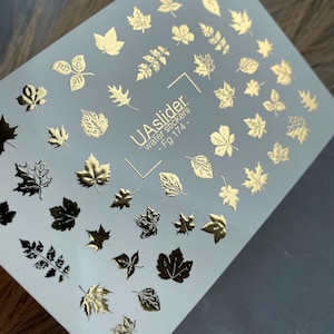May include: A white sheet of nail art water stickers featuring various gold metallic leaf designs. The center of the sheet displays the text UAslider water stickers - Fg 174. The sheet contains multiple individual leaf shapes in different sizes.