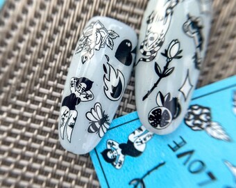 May include: Two clear nail polish stickers with black and white designs. The designs include a rose, a heart, a flame, a bee, a pin-up girl, and a snake. The stickers are on a gray textured surface.