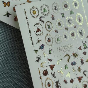 Może przedstawiać: Dwa arkusze naklejek wodnych do zdobienia paznokci z różnymi owadami, w tym chrząszczami, motylami i ważkami. Niektóre owady są oprawione w owalne, złote ramki. Widoczny jest tekst "UAslider water stickers - CF 788".