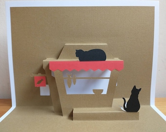 May include: A pop-up card with a brown background and a red awning. Two black cats are on the card, one is sitting and the other is lying down. The card has a fish cutout on the side.