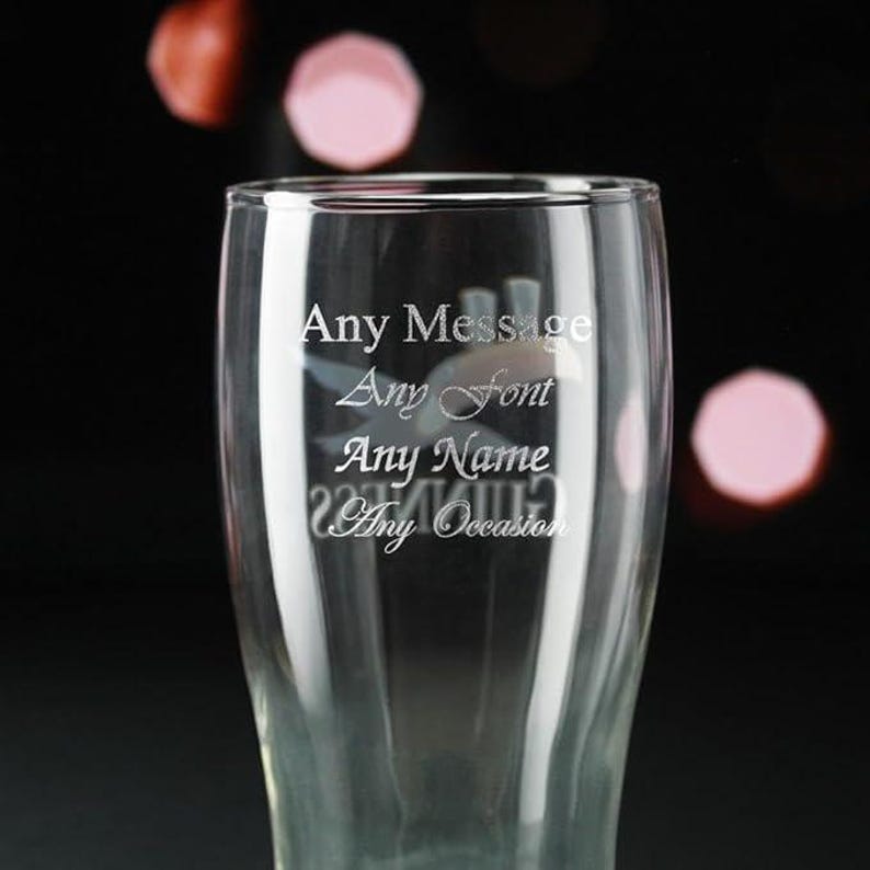 May include: A clear pint glass with engraved text. The text reads "Any Message, Any Font, Any Name, Any Occasion." The glass has a curved shape and is set against a dark background with blurred pink and red highlights.