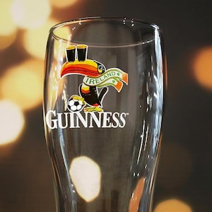 Może przedstawiać: Przezroczysta szklanka do piwa z białym logo Guinness oraz kolorową grafiką tukana balansującego dwoma szklankami na dziobie, trzymającego piłkę nożną i noszącego szalik z napisem Ireland oraz małymi zielonymi koniczynami.