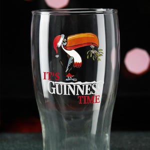 Puede incluir: Un vaso de pinta Guinness transparente con un tucán festivo. El tucán lleva un gorro de Papá Noel y sostiene muérdago. Las palabras "IT'S GUINNESS TIME" están impresas en el vaso. El vaso tiene forma curva.