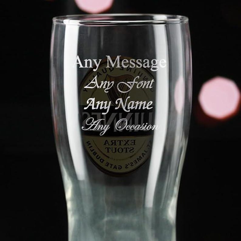 May include: A clear pint glass with the words "Any Message, Any Font, Any Name, Any Occasion" in white script. The glass has a logo in the center. The background is dark with soft pink orbs.