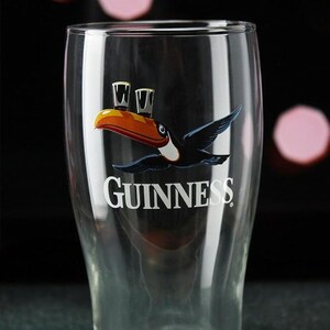May include: A clear pint glass with the Guinness logo. The logo features a toucan carrying a pint of Guinness, and the word "Guinness" in white. The glass is tapered and has a wide mouth, typical of a pint glass.