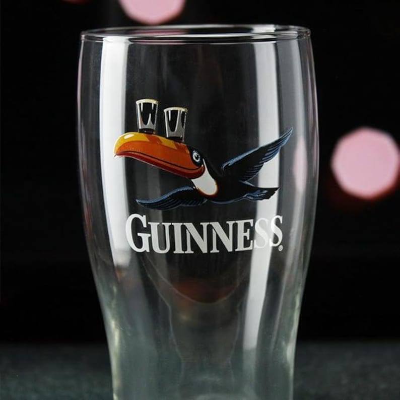 May include: A clear pint glass with the Guinness logo. The logo features a toucan carrying a pint of Guinness, and the word "Guinness" in white. The glass is tapered and has a wide mouth, typical of a pint glass.