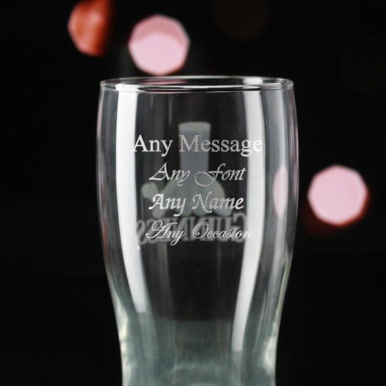 May include: A clear glass pint glass with the words "Any Message, Any Font, Any Name, Any Occasion" etched on the front. The glass has a curved shape and is set against a blurred background with pink and red bokeh.