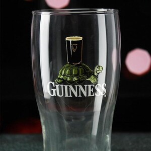 May include: A clear pint glass with the Guinness logo. The logo features a black pint of Guinness atop a green turtle, with the word "Guinness" in white below. The glass is empty.