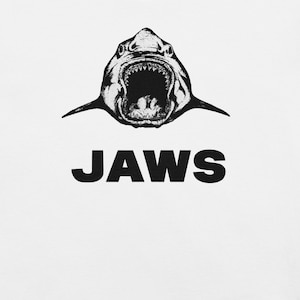 Puede incluir: Gráfico en blanco y negro de un tiburón con la boca abierta, dientes visibles, y la palabra "JAWS" en letras negras en negrita debajo. La imagen está sobre un fondo blanco.