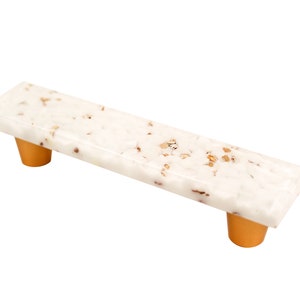 Puede incluir: Tirador de armario rectangular blanco con detalles dorados. El tirador tiene un aspecto moteado, marmoleado.