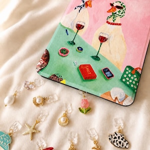 Könnte beinhalten: Eine rechteckige Tablethülle mit einem Gemälde von zwei Enten, die Kopftücher und Sonnenbrillen tragen und an einem grünen Tisch Wein trinken, neben einem roten Buch mit der Aufschrift SASSY. Darunter befinden sich verschiedene kleine Metallanhänger, darunter eine rosa Tulpe, Seesterne, Muscheln und ein Hut mit Kuhfleckenmuster.