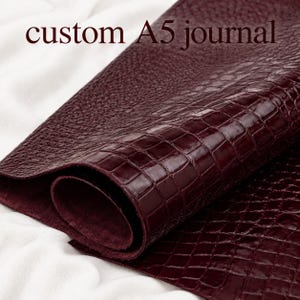 May include: A deep burgundy A5 journal cover featuring a glossy, embossed crocodile skin texture on the exterior and a matte, suede-like finish on the interior. The text custom A5 journal is printed in a dark font at the top of the image.