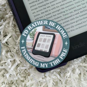 May include: A round sticker with the text "I'd Rather Be Home Finishing My TBR Pile" encircling an illustration of an e-reader with the words "1000 Books" on the screen. The sticker is placed on a Kindle e-reader.