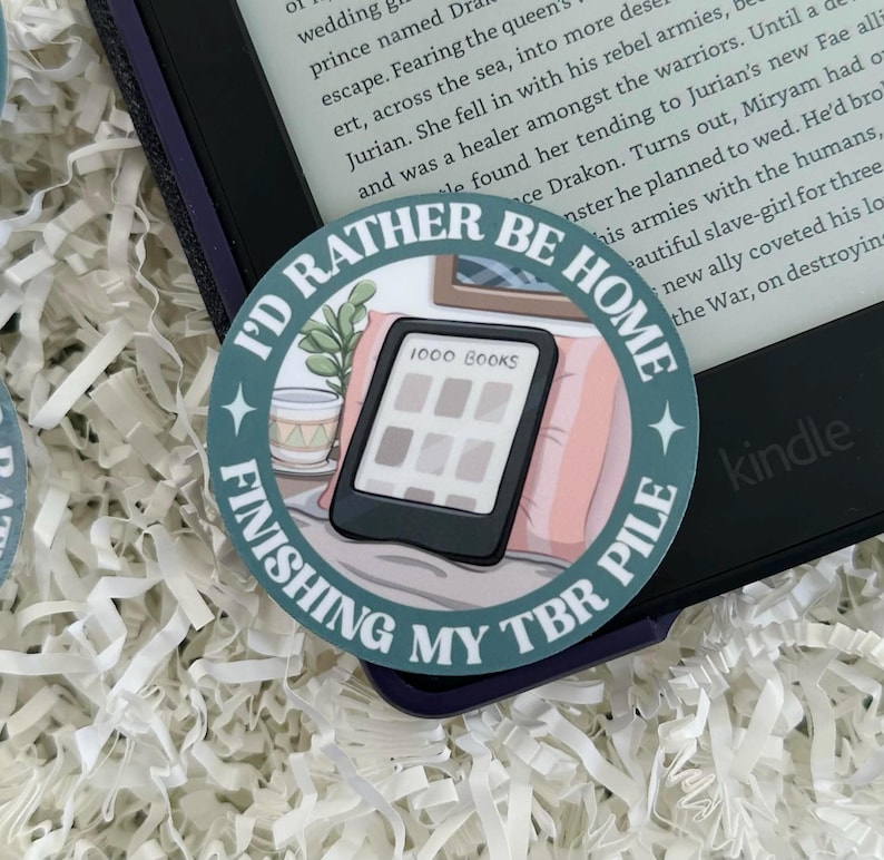 May include: A round sticker with the text "I'd Rather Be Home Finishing My TBR Pile" encircling an illustration of an e-reader with the words "1000 Books" on the screen. The sticker is placed on a Kindle e-reader.