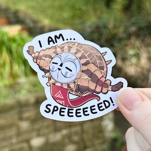 May include: A sticker depicting a cartoon owl-like character with the words "I AM... SPEEEEEEED!". The creature has a round face, large eyes, and a shell-like body with brown and tan details. A red banner with a symbol is held in its hands.