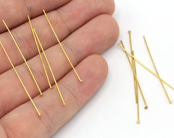 Pode incluir: Um close-up de um grupo de alfinetes de cabeça dourada. Os alfinetes medem aproximadamente 2,5 cm de comprimento e têm uma cabeça arredondada. Eles estão dispostos em um padrão aleatório em uma superfície branca.