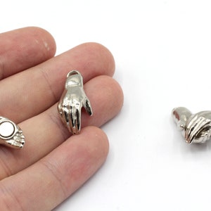 Op de afbeelding: Drie zilveren metalen handcharms. Twee van de charms zijn enkele handen met de palm naar boven. De derde charm is twee handen die in elkaar grijpen.