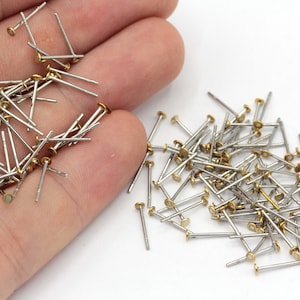 May include: A collection of small, silver-colored metal pins with gold-colored flat heads. The pins are scattered on a white surface, with some held in a hand. These are likely jewelry-making or craft supplies.