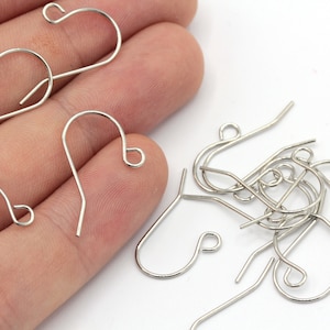 May include: Silver-toned earring hooks, a group of hooks are scattered on a white surface, and a hand is holding three hooks.