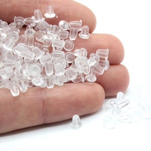 May include: A pile of clear plastic earring backs in a hand. The backs are small and round with a small post sticking out of the top.