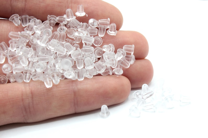 May include: A pile of clear plastic earring backs in a hand. The backs are small and round with a small post sticking out of the top.