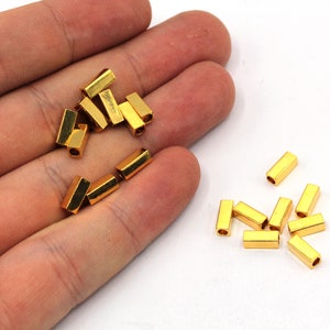 Op de afbeelding: Een stapel gouden metalen buisspacers voor het maken van sieraden. De spacers zijn ongeveer 1,2 cm lang en hebben een gladde, glanzende afwerking.