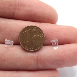 May include: Two clear plastic earring backs next to a 1 Euro coin. The earring backs are small and cylindrical.
