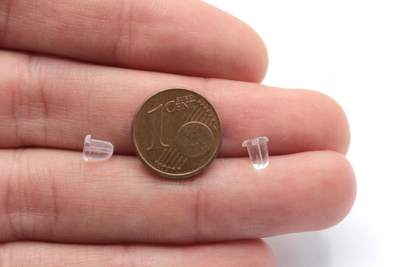 May include: Two clear plastic earring backs next to a 1 Euro coin. The earring backs are small and cylindrical.