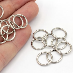 May include: A pile of silver jump rings, also known as key rings, are shown on a white surface. The rings are small and round, and they are perfect for attaching to jewelry, keychains, or other crafts.