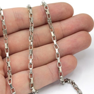 May include: A silver chain necklace with a box chain design. The chain is lying on a hand.