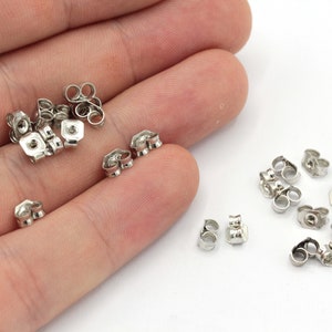 May include: A close-up of a hand holding a pile of silver earring backs. There are also a few earring backs scattered on a white surface.