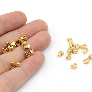 May include: A close-up of a pile of gold heart-shaped beads. The beads are small and have a shiny finish.