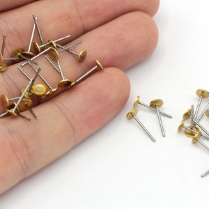 May include: A pile of gold-colored metal earring posts with silver stems. The earring posts are scattered on a white surface.