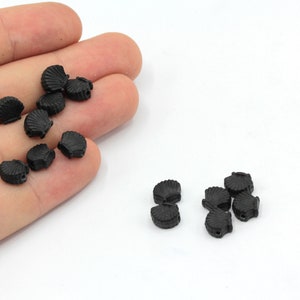 Può includere: Una collezione di piccole perline nere a forma di conchiglia. Le perline sono realizzate in un materiale solido e hanno una superficie dettagliata e strutturata. Sono sparse su uno sfondo bianco, suggerendo il loro utilizzo in progetti di gioielleria o artigianato.