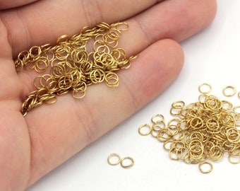 Puede incluir: Un montón grande de anillos de salto dorados, un suministro popular para la fabricación de joyas, se dispersa sobre una superficie blanca. También hay algunos montones más pequeños de anillos de salto.