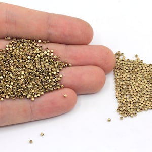 Könnte beinhalten: Ein großer Haufen goldfarbener Metallperlen in einer Hand und ein kleinerer Haufen der gleichen Perlen auf einer weißen Oberfläche.