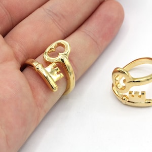 May include: Two gold-toned key-shaped rings. The rings are designed to look like an old-fashioned skeleton key. One ring is worn on a finger, the other is lying flat.