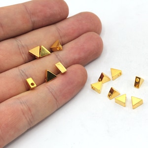 Puede incluir: Una colección de cuentas de metal de forma triangular de color dorado con un agujero en el centro. Las cuentas están esparcidas sobre una superficie blanca.