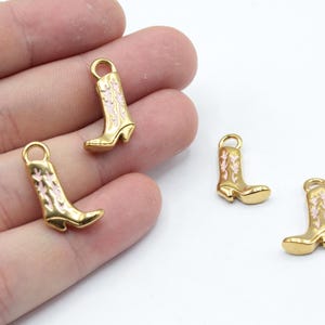 May include: Three gold-coloured boot charms with pink floral designs. The charms have a loop at the top for attaching to a bracelet or necklace.