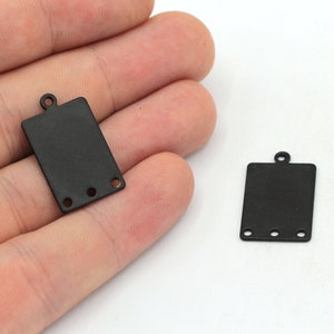 May include: Two black metal rectangular tag charms with three holes each. The charms have a loop at the top for attaching to a necklace or bracelet.